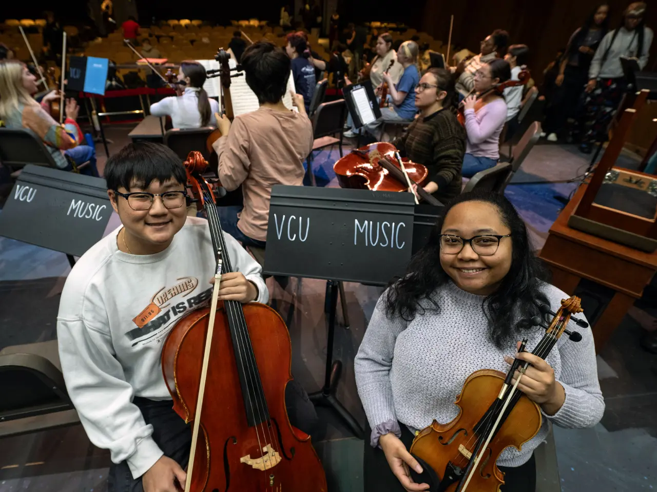 Two students stand by a music stand with their string instruments