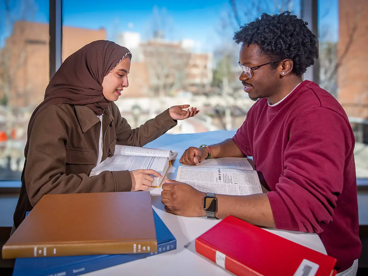 Two students sit together surrounded by books on a table