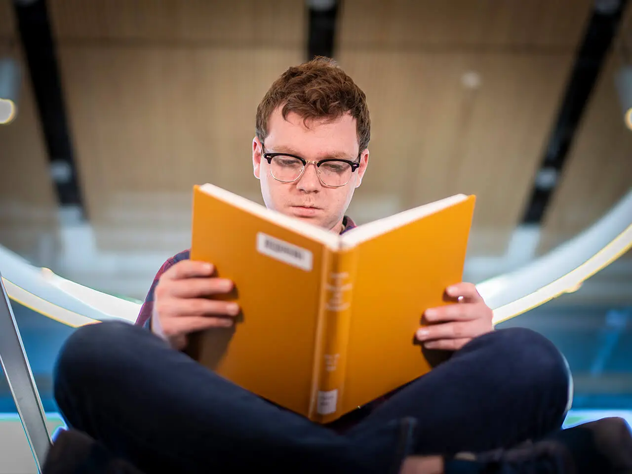 A student wearing glasses while reading a gold colored text book