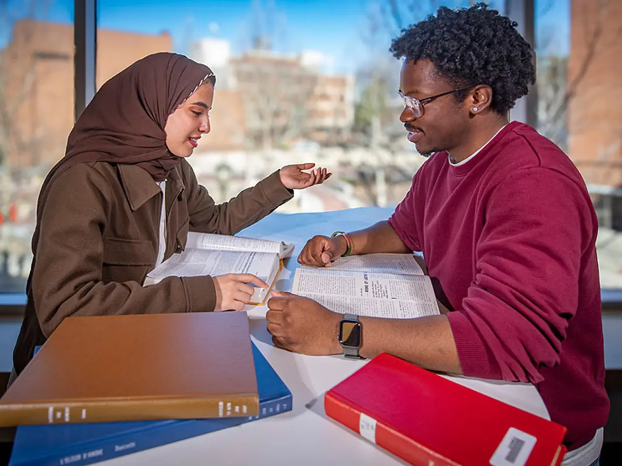 Two students sit together surrounded by books on a table